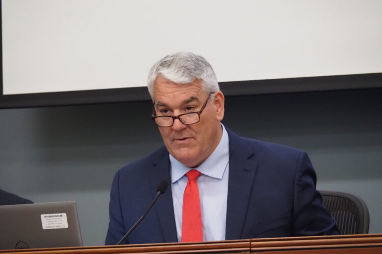 Andy Zay leads a meeting. He is a white man with glasses, white hair, and wearing a blue suit with a red tie.
