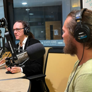 Graham Watson and Braiden Robinson sit in a radio studio wearing headphones. Graham is talking into a microphone.