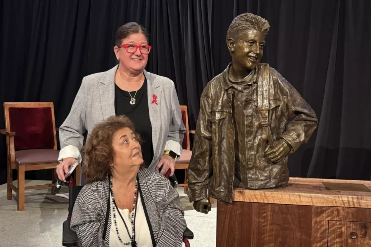A bronze sculpture of Ryan White was unveiled at the Indiana Memorial Union. White's mom, Jeanne White-Ginder, and his sister, Andrea White, stand next to it. The bronze statue is from the waist up and is placed on a wooden pedestal.