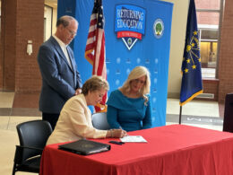 U.S. Education Secretary Linda McMahon sits to sign a state waiver on education funding. She is joined at the table by state Education Secretary Katie Jenner. Standing behind them is Indiana Gov. Mike Braun.