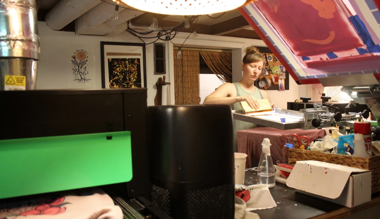 A woman stands in a small cluttered room. She operates a machine that prints designs onto clothing