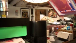 A woman stands in a small cluttered room. She operates a machine that prints designs onto clothing