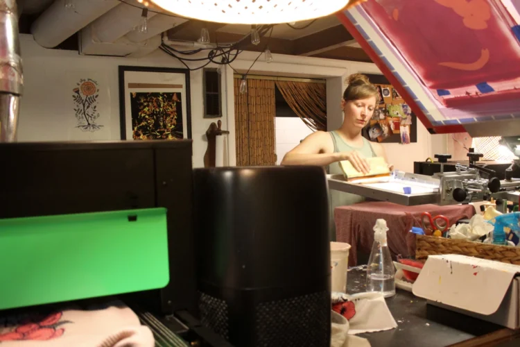 A woman stands in a small cluttered room. She operates a machine that prints designs onto clothing