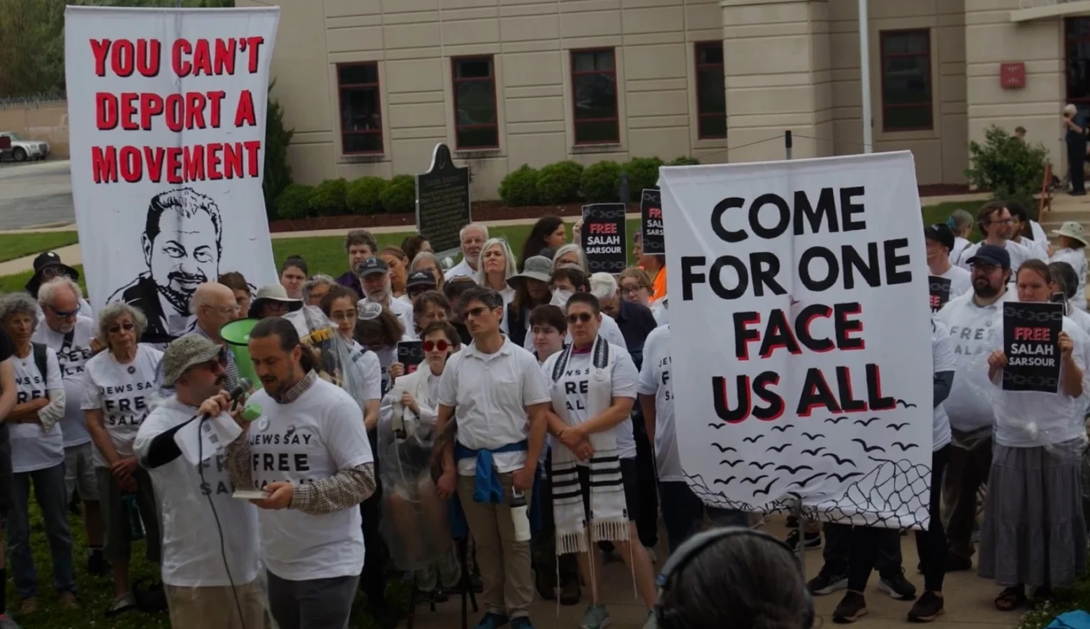 A crowd of protestors in white shirts stand outside with signs. A man in front of the crowd holds a phone and speaks into a microphone