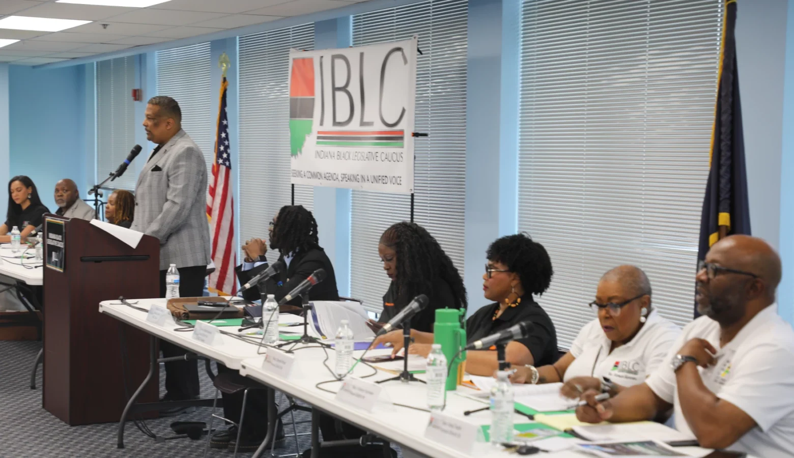 Members of the Indiana Black Legislative Caucus sit at long tables and stand at a podium at the front of a room, speaking to constituents.
