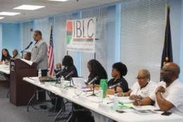 Members of the Indiana Black Legislative Caucus sit at long tables and stand at a podium at the front of a room, speaking to constituents.