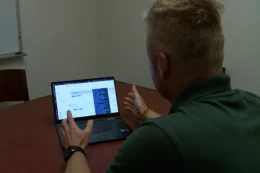 A man, seen from behind, sits at a table in front of a laptop computer. He gestures to the screen.