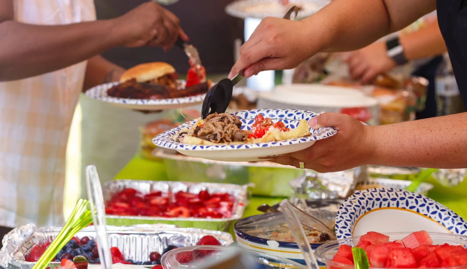 Hands hold plates and fill them with food from a buffet table, including pulled pork, watermelon cubes, and berry salad.