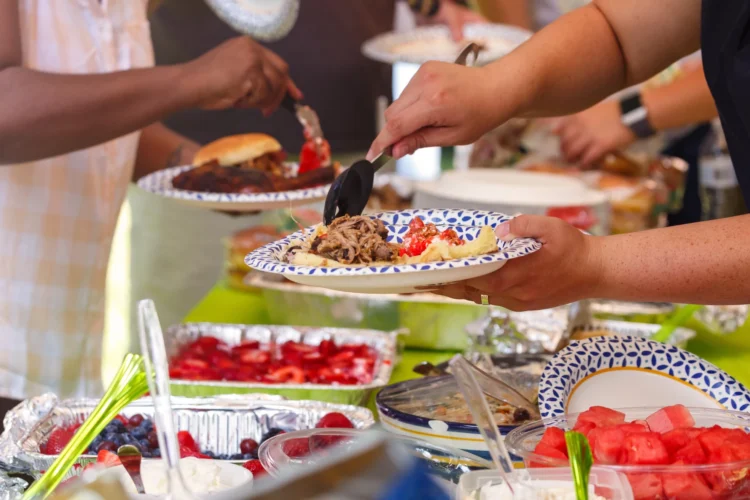 Hands hold plates and fill them with food from a buffet table, including pulled pork, watermelon cubes, and berry salad.