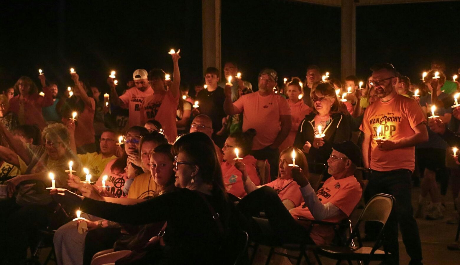 Many people stand together, wearing pink t-shirts and holding lit candles.