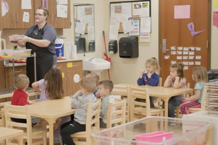 A teacher in a childcare classroom is at the front of the room taking what appears to be a craft object from a cart in front of eight children of kindergarten age seated at desks.