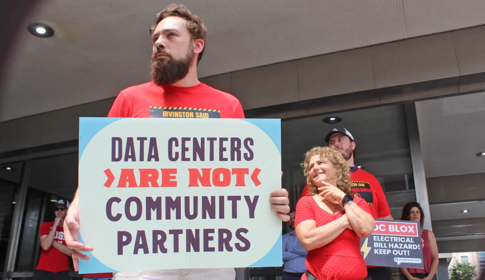 Red-shirted protesters are holding signs saying, among other messages, "Data Centers Are Not Community Partners." Protesters are seen standing outside of the Indianapolis-Marion County city county building.