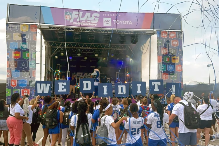 Players and supporters celebrate reaching 100 girls flag football teams statewide — the threshold required for Indiana High School Athletic Association sanctioning — during the NFL FLAG Championships on Sunday, July 26, 2026, in Westfield, Ind. An elevated stage at the Westfield sports facility displays flag football logos and the NFL Flag Football Championship logo above the stage. On the stage is the Colts mascot. In front of the stage, supporters are holding individual letter signs with white letters on a blue background spelling out "WE DID IT." Dozens of supporters are in front of the stage celebrating with streamers flying through the air.