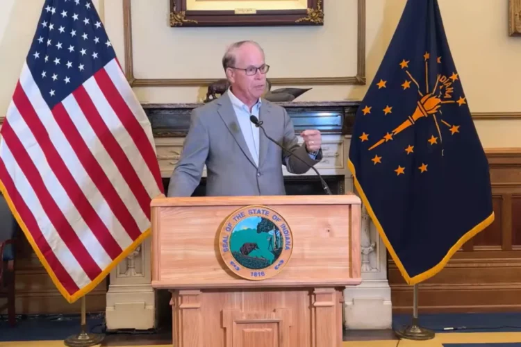 Gov. Braun, wearing a gray jacket and white shirt without a tie, stands behind a wooden podium with a microphone and is flanked by the American flag on the left of the frame, and the Indiana flag on the right. The flags are hanging downward and displayed on vertical poles approximately eight feet tall.