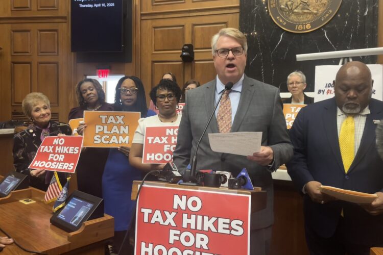 Indiana Democrats stand on the House floor at a press conference, holding red signs that read No Tax Hikes For Hoosiers and Tax Plan Scam.