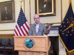 Governor Mike Braun speaks from a podium in his office, surrounded by the American and Indiana flags.