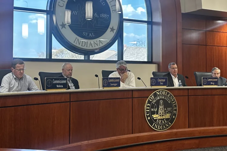 Members of the Indiana Utility Regulatory Commission listen to customers' concerns about utility rates during public meeting at Noblesville City Hall, Thursday, April 9, 2026. Five commissioners are seated at a curved bench behind wood paneling with name signs and microphones in front o them. A large Noblesville city seal is on an illuminated wall behind the commissioners.