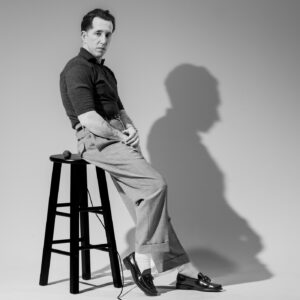 A promotional photo where Pokey LaFarge sits on the edge of a stool. There is a wired microphone next to him on the seat of the stool.