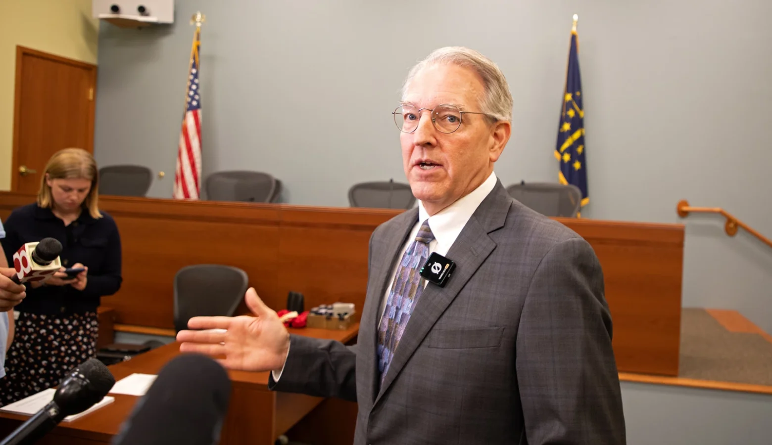 Indiana State Rep. Matt Pierce (D-Bloomington) held a press conference after the IURC’s updates on utility investigations Wednesday, July 15, 2026. He is standing while talking to reporters. He is wearing a gray suit and gesturing with his right hand. His press conference was held in a council meeting room with a panel with chairs (unoccupied) in the background, and other chairs and tables in the foreground where reporters are standing.