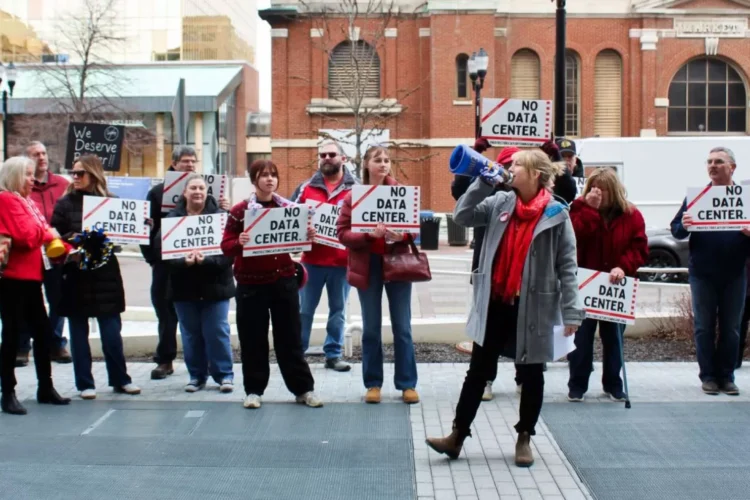 Many people stand outside, holding signs that read No Data Center. An organizer stands in front, talking through a bullhorn.