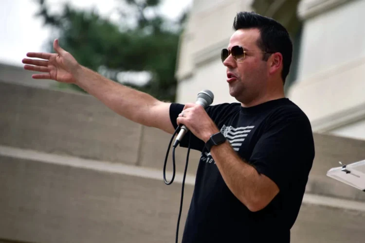 Lieutenant Governor Micah Beckwith speaks at a rally otudoors, wearing a t-shirt with an American flag and sunglasses. He holds a microphone.