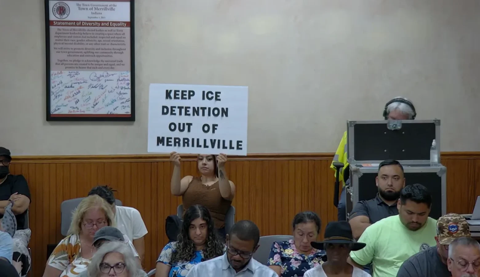 A woman holds up a protest sign at the back of a town council meeting that reads Keep ICE Detention Out of Merrillville.