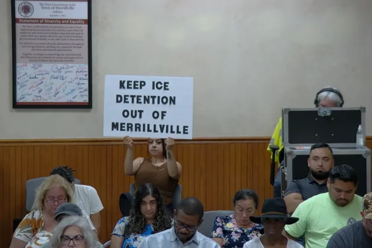 A woman holds up a protest sign at the back of a town council meeting that reads Keep ICE Detention Out of Merrillville.