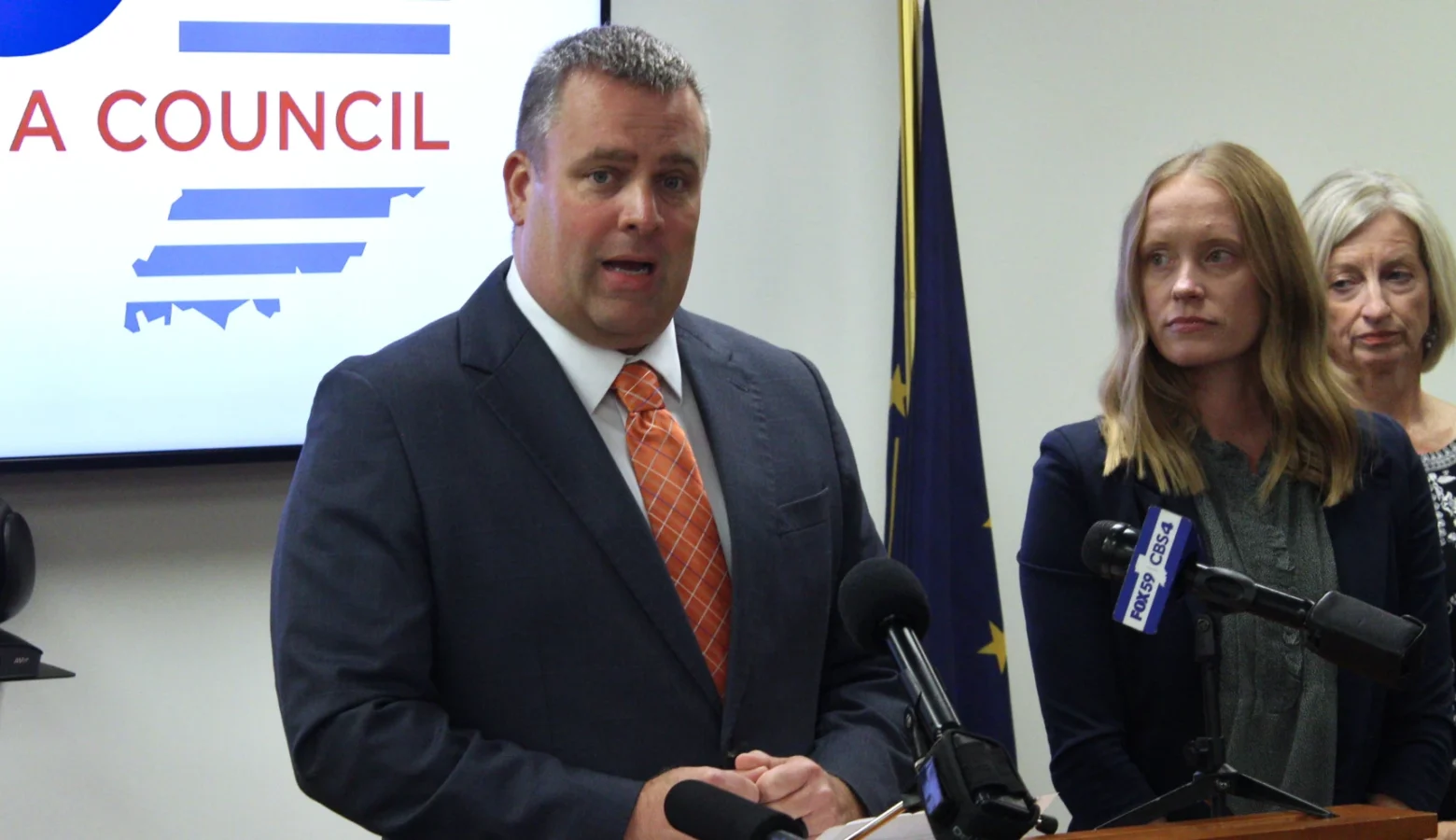 Brian Gould, executive director of the Build Indiana Council, is calling for lawmakers to address a road funding cliff the state is headed towards. In the photo he is standing in front of a podium with microphones. He is wearing a gray suit with an orange tie. Two women, presumably staff members of his organization, are in the right of the frame.