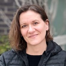 Ann Larson is a white woman with brown eyes and shoulder-length brown hair parted at the side. She is wearing a black shirt covered by a black jacket.