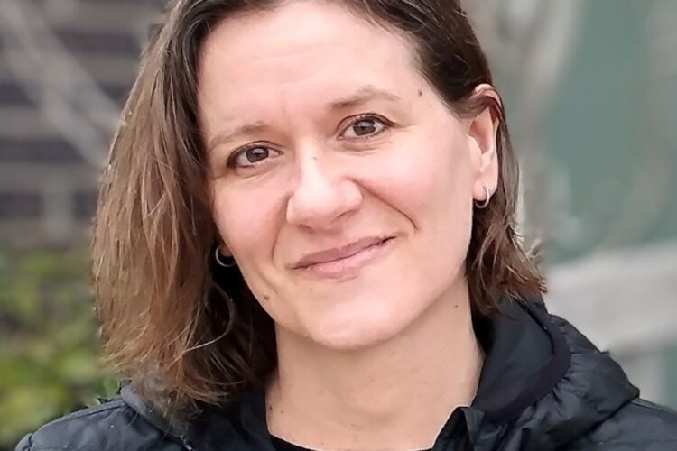 Ann Larson is a white woman with brown eyes and shoulder-length brown hair parted at the side. She is wearing a black shirt covered by a black jacket.