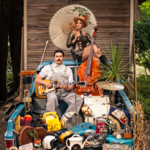 Two people sit with instruments in a truck bed filled with items. A.W. Cardinal holds a small stringed instrument, while Jasmine Colette sits on top of the cab holding a parasol and an upright base.