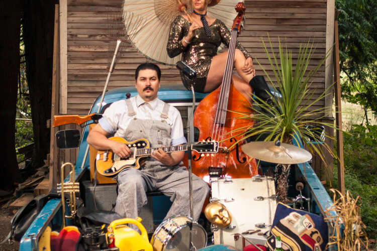 Two people sit with instruments in a truck bed filled with items. A.W. Cardinal holds a small stringed instrument, while Jasmine Colette sits on top of the cab holding a parasol and an upright base.