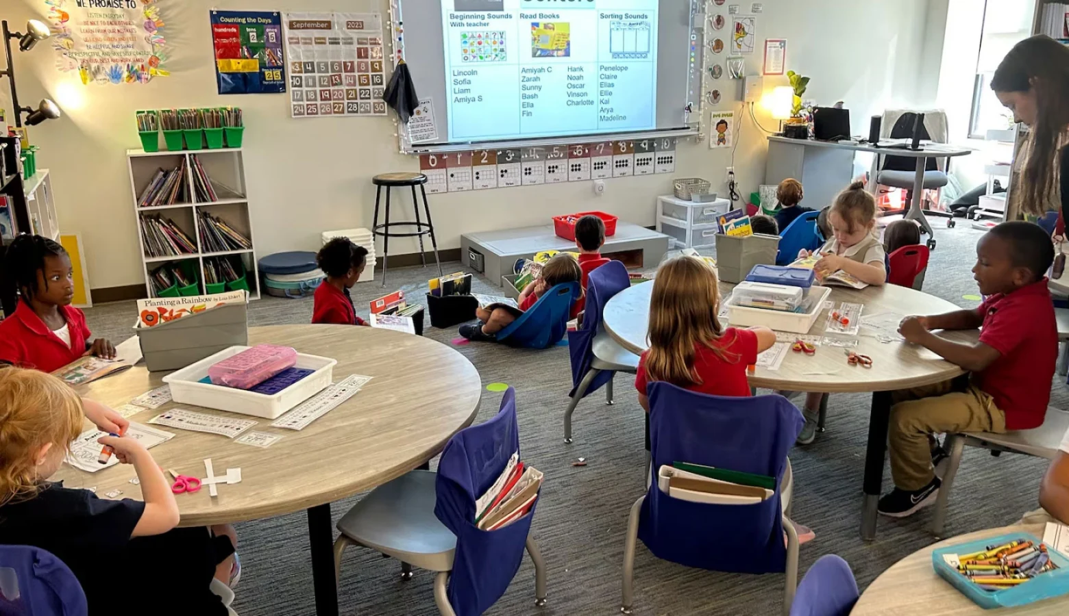 Kindergartners in class at Herron Prep Academy in Indianapolis in September 2023. 11 students and a teacher are seated around round tables and the floor looking at a projected slide on a white board. Art supplies like crayons and various papers are also in the photo.