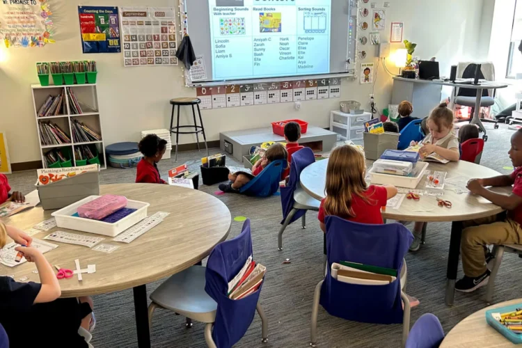 Kindergartners in class at Herron Prep Academy in Indianapolis in September 2023. 11 students and a teacher are seated around round tables and the floor looking at a projected slide on a white board. Art supplies like crayons and various papers are also in the photo.