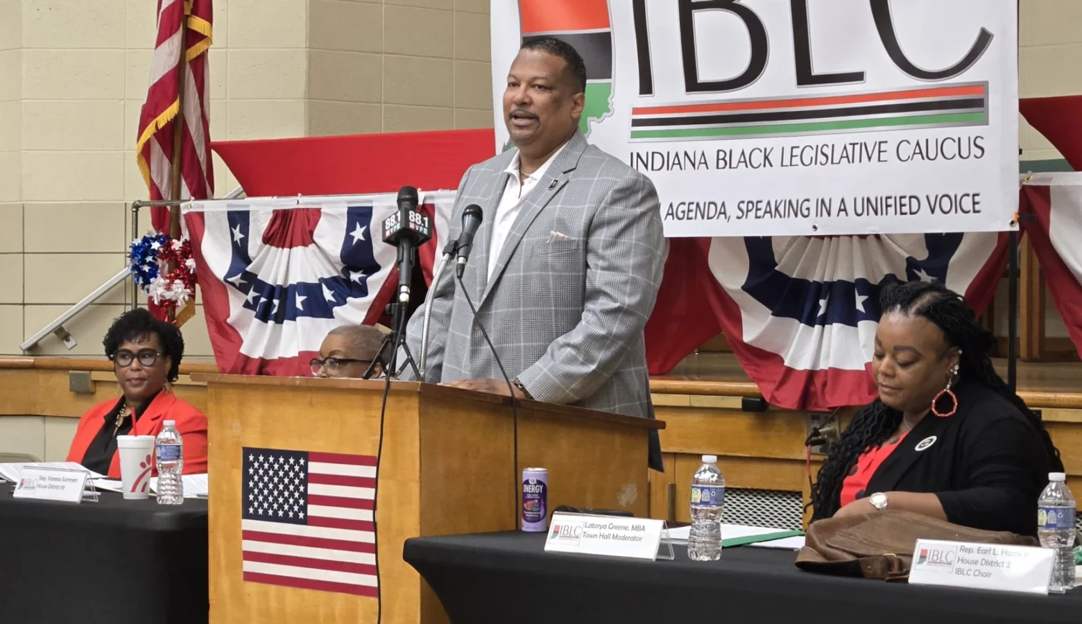Indiana Black Legislative Caucus Chair Earl Harris Jr. (D-East Chicago) addresses a gathering from a podium equipped with microphones and an American flag on the front.