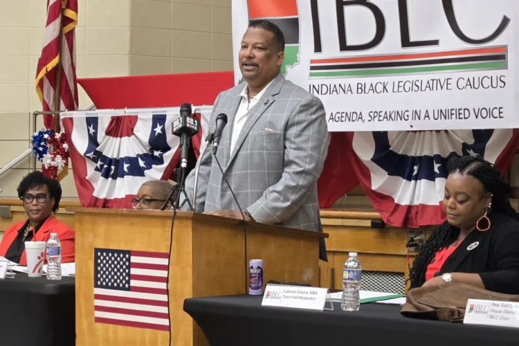 Indiana Black Legislative Caucus Chair Earl Harris Jr. (D-East Chicago) addresses a gathering from a podium equipped with microphones and an American flag on the front.