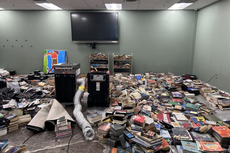 A large room with thousands of damaged books stacked into piles on top of plastic tarps. In the center of the room is two square machines, one of them has a metal tube attached to it.