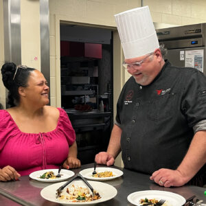 Kara DuQuette and Chef Jason stand smiling behind a metal table with multiple servings of salad. Chef Jason Reynolds wears a high chef's hat.