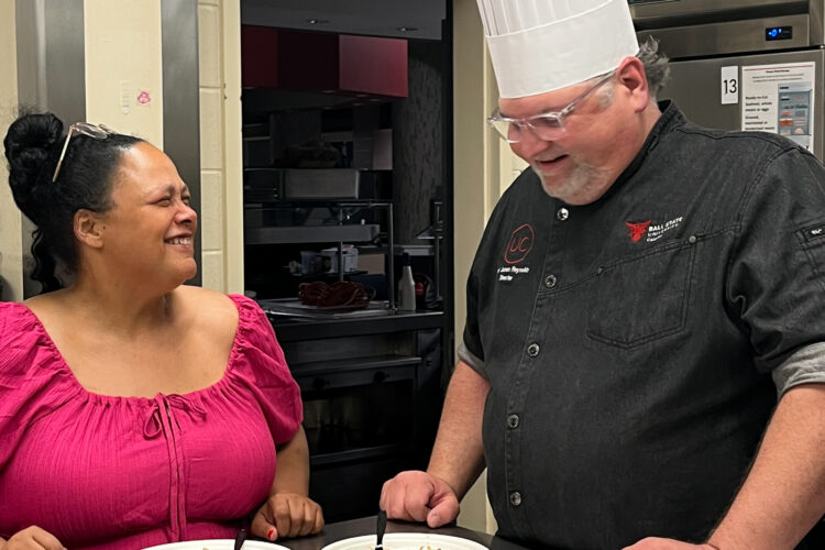 Kara DuQuette and Chef Jason stand smiling behind a metal table with multiple servings of salad. Chef Jason Reynolds wears a high chef's hat.