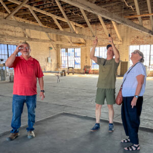 Jason and Jerrad Oakley stand with Michelle Kinsey; all three of them are in an industrial building, looking up at the rafters. It's very bright from natural sunlight.