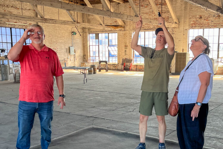 Jason and Jerrad Oakley stand with Michelle Kinsey; all three of them are in an industrial building, looking up at the rafters. It's very bright from natural sunlight.