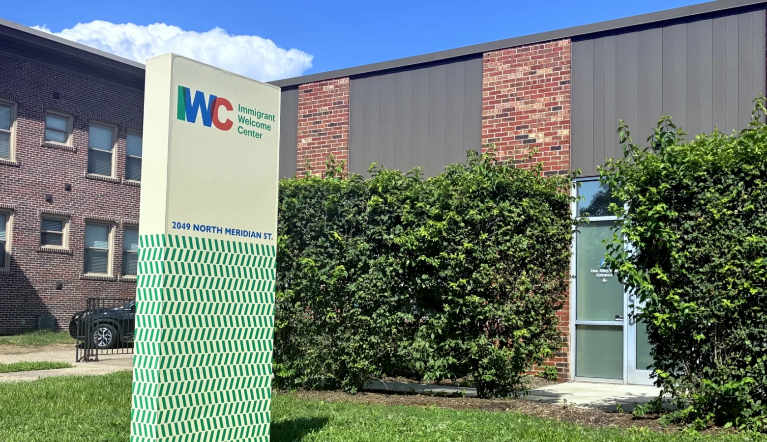 The photo shows an exterior shot of the IWC (Immigrant Welcome Center) whose sign is a vertical rectangle with the IWC logo at the top and green forward and back slashes below. It stands in front of a two-story brick building. The sky above it is blue with a fair weather cumulus cloud.
