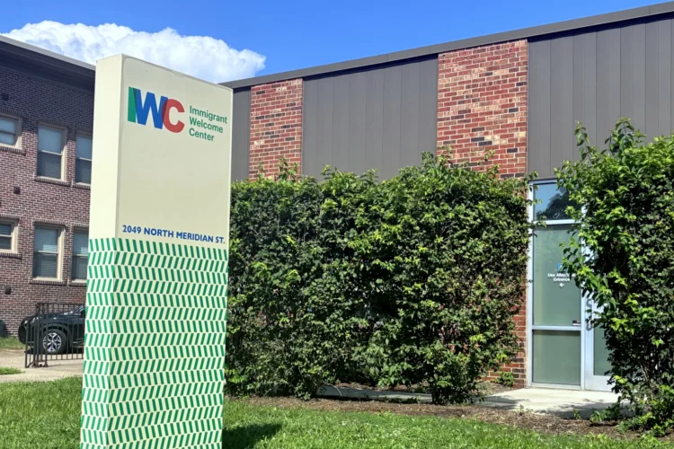 The photo shows an exterior shot of the IWC (Immigrant Welcome Center) whose sign is a vertical rectangle with the IWC logo at the top and green forward and back slashes below. It stands in front of a two-story brick building. The sky above it is blue with a fair weather cumulus cloud.