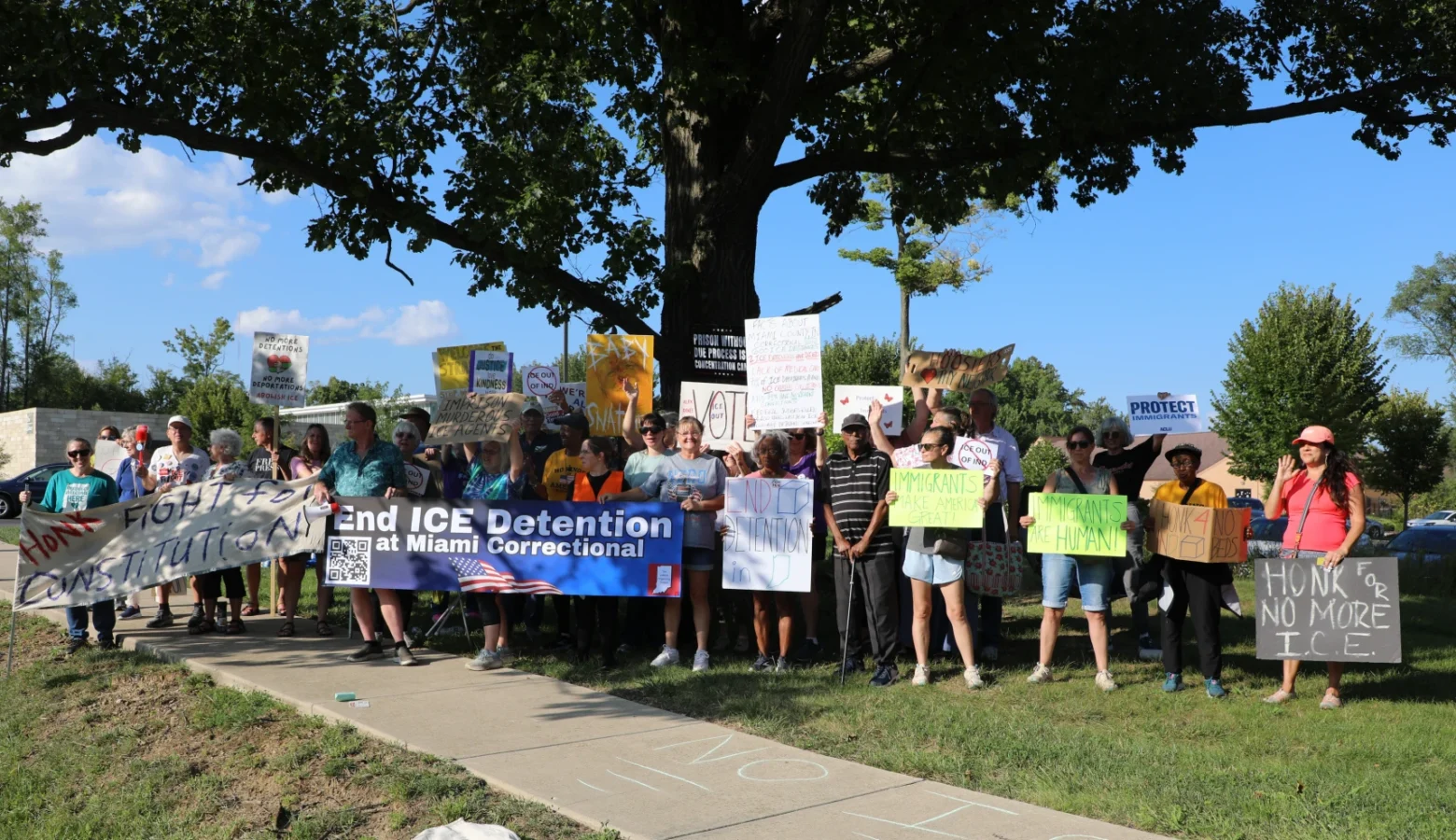 A group of around two dozen protesters hold signs saying ICE detention should end at Miami Correctional. They are standing under a large tree near a road on a sunny day.