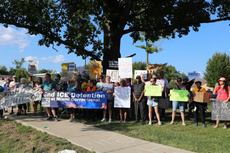 A group of around two dozen protesters hold signs saying ICE detention should end at Miami Correctional. They are standing under a large tree near a road on a sunny day.