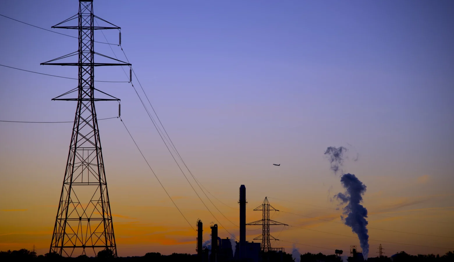 A photo of a high-voltage utility tower with power lines leading from a power generating station with a smokestack emitting steam into a clear sky at sunset.