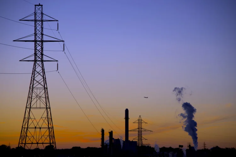 A photo of a high-voltage utility tower with power lines leading from a power generating station with a smokestack emitting steam into a clear sky at sunset.