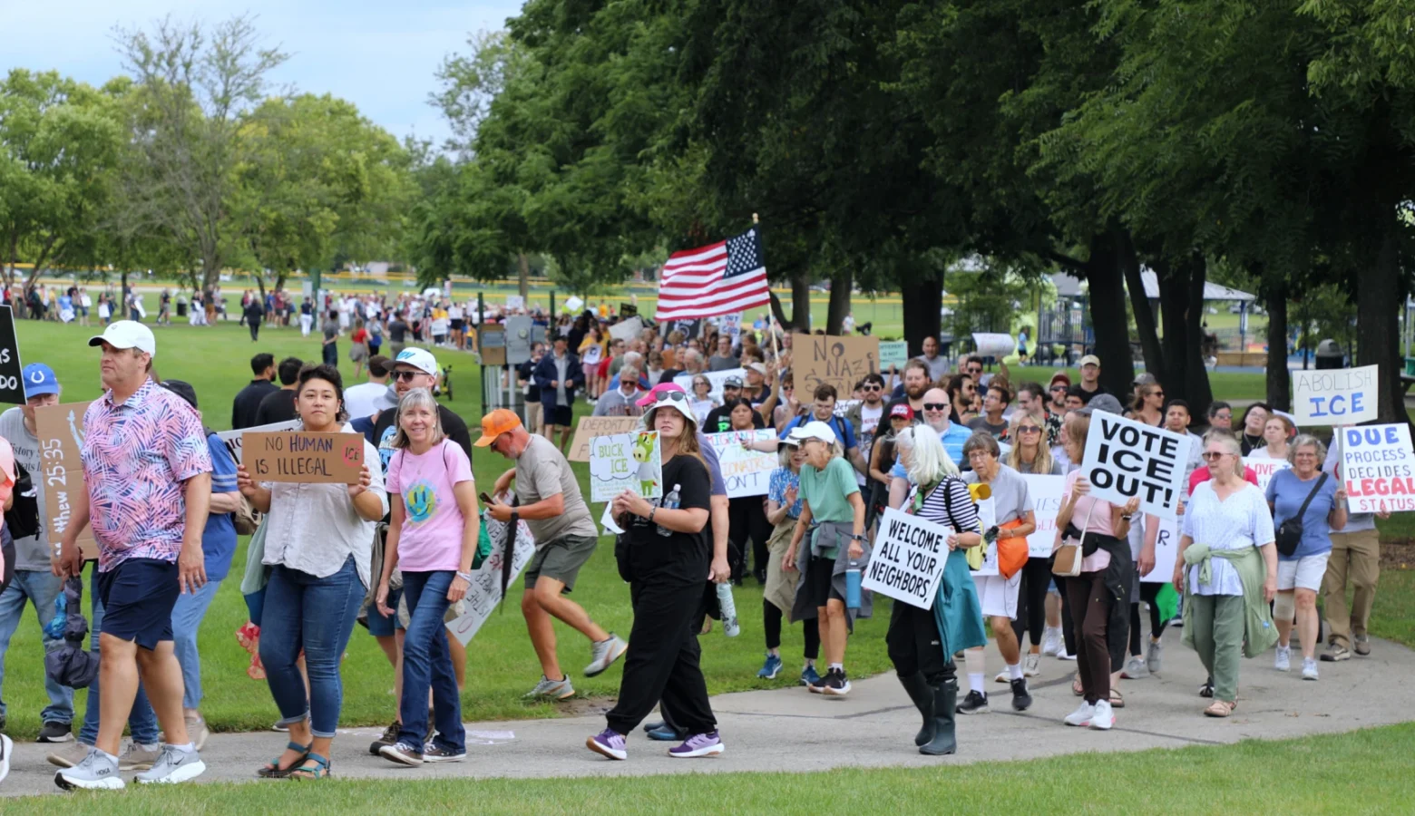 Hundreds marched in support of immigrants Aug. 1, 2026 in Fishers, largely framed as a counter-event to a pro-mass deportation rally happening nearby. The marchers are carrying signs and American flag and are walking on a sidewalk that winds through trees.