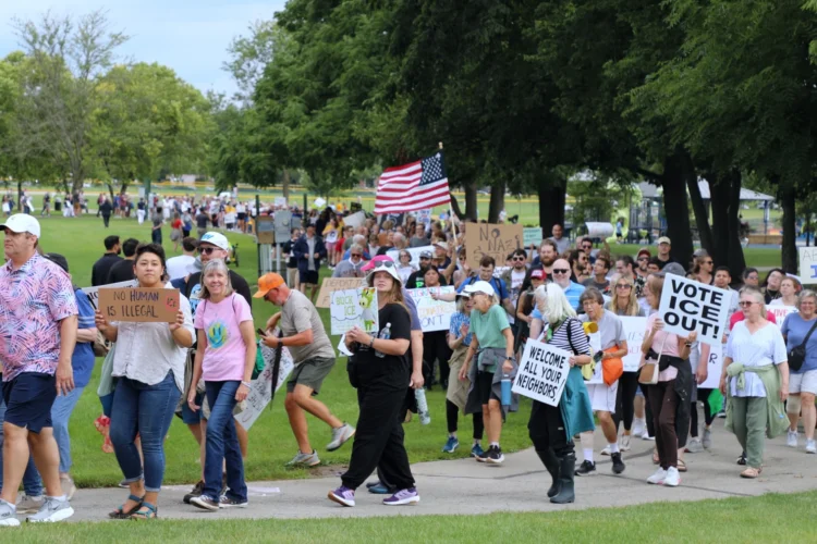 Hundreds marched in support of immigrants Aug. 1, 2026 in Fishers, largely framed as a counter-event to a pro-mass deportation rally happening nearby. The marchers are carrying signs and American flag and are walking on a sidewalk that winds through trees.