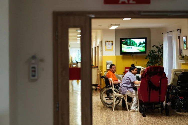 The photo shows a wide shot of a psychiatric ward with a staff member seated at a table with what appears to be three patients.